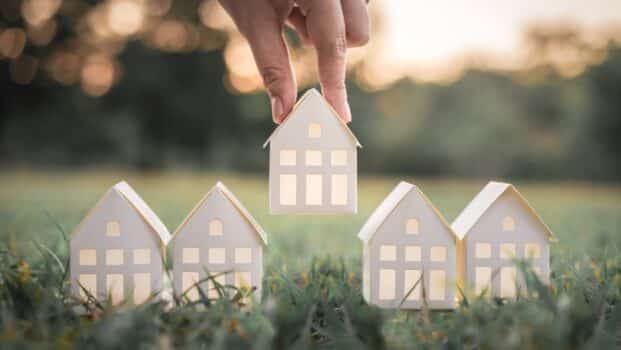 Hand choosing white paper house model from group of house on green grass, selective focus, planning to buy property. Choose what's the best. A symbol for construction ,ecology, loan concepts