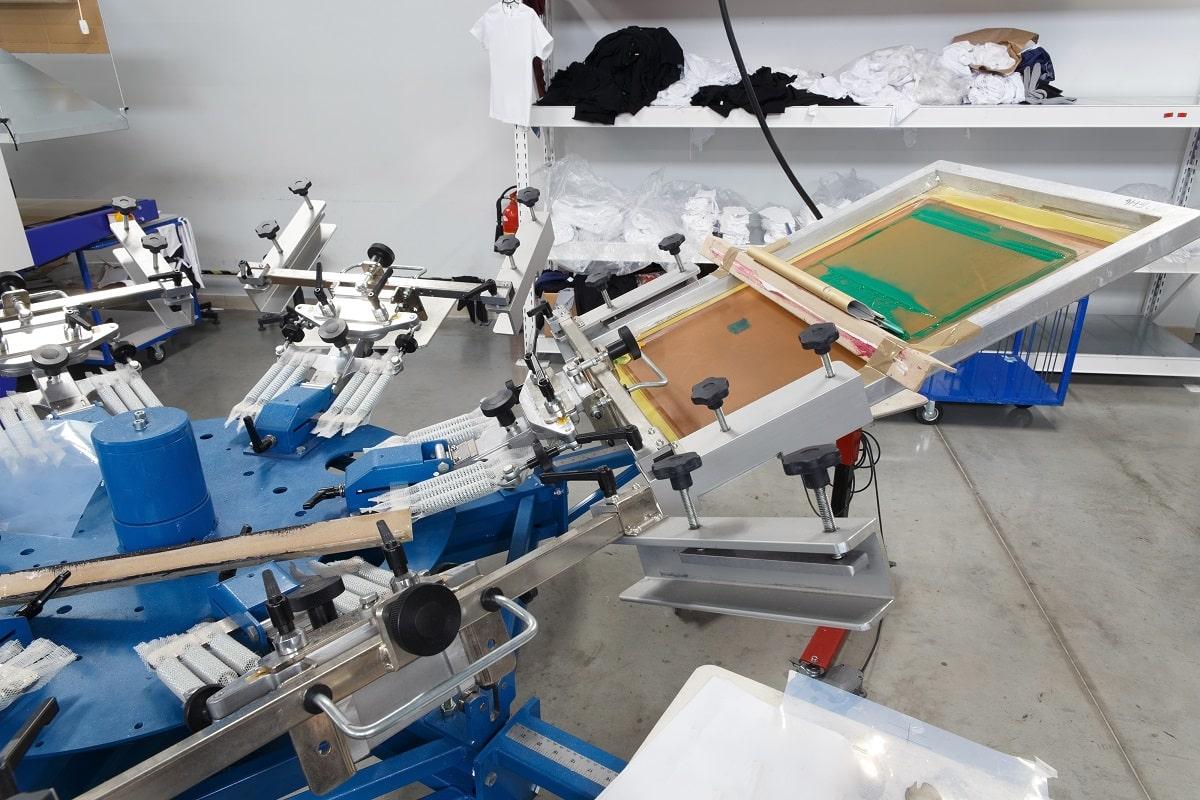 Screen printing machine for applying images by silkscreen printing. Print screening apparatus. Serigraphy production. Printing images on t-shirts in a design studio. How to do screen printing.