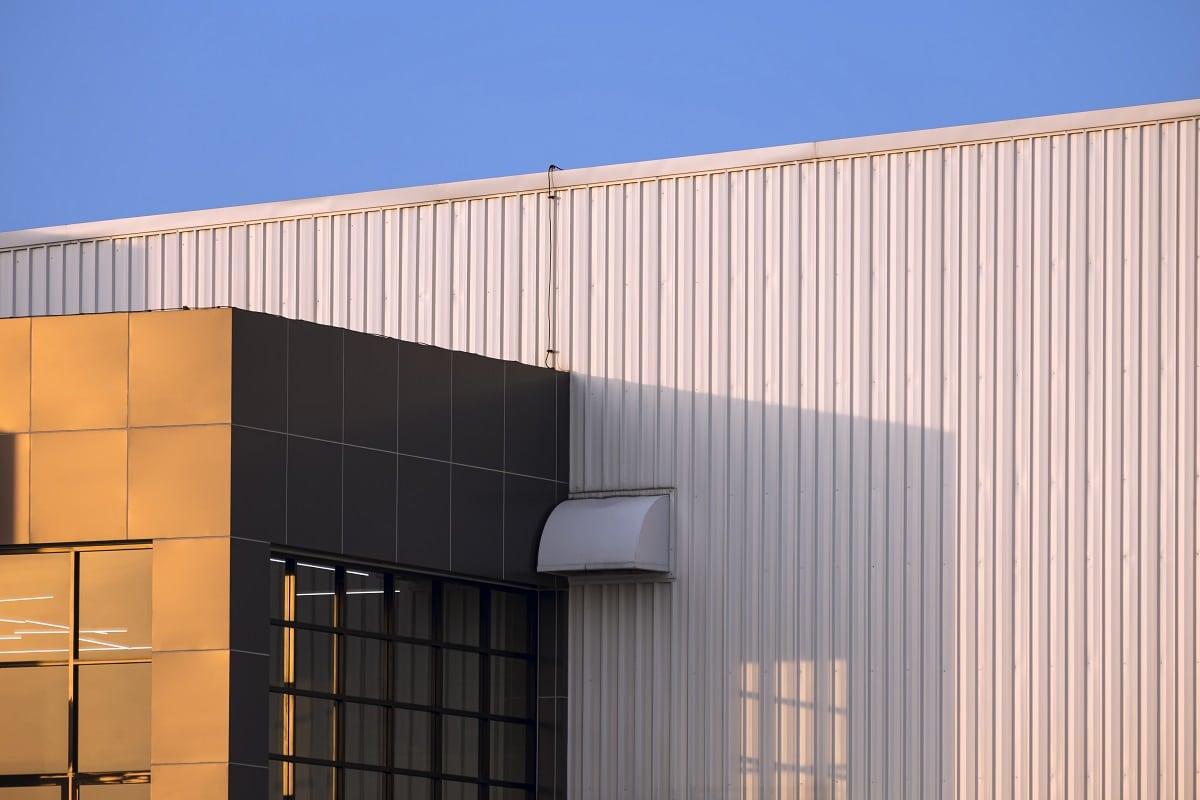 Sunlight on part of brown modern office on white corrugated metal wall of industrial building against blue sky background in evening time. Seamless steel log siding.